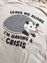 Load image into Gallery viewer, T-shirt with an opossum graphic and text 'Leave me alone I'm having a crisis' on a beige background.